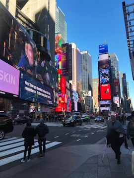 A bustling urban scene with numerous large digital billboards displaying various advertisements. Tall buildings surround the area, and people walk across the crosswalk. Vehicles are visible on the street, contributing to the lively city atmosphere under a clear blue sky.
