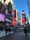 A bustling urban scene with numerous large digital billboards displaying various advertisements. Tall buildings surround the area, and people walk across the crosswalk. Vehicles are visible on the street, contributing to the lively city atmosphere under a clear blue sky.