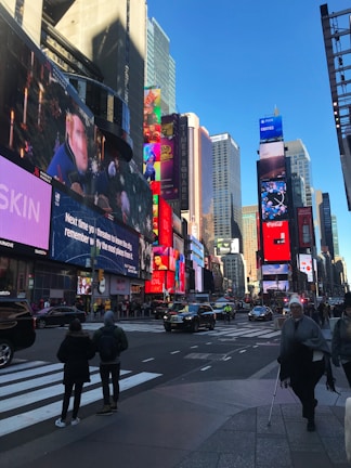 A bustling urban scene with numerous large digital billboards displaying various advertisements. Tall buildings surround the area, and people walk across the crosswalk. Vehicles are visible on the street, contributing to the lively city atmosphere under a clear blue sky.