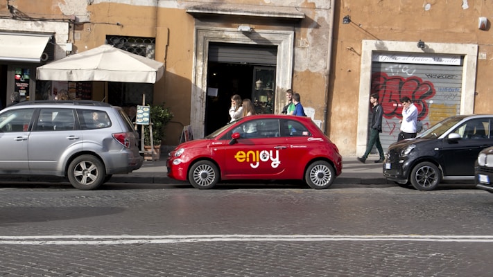 A row of parked cars along a street in an urban area, with a notable red compact car in the center displaying the word 'enjoy'. The cars are lined up against a background of a rustic building facade with large windows and graffiti. People are walking on the sidewalk alongside the cars, some appearing to be engaged in conversation.