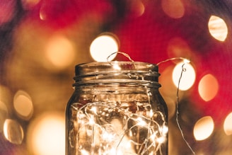 A glass jar filled with small LED fairy lights is placed in front of a blurred background of circular bokeh lights. The jar's transparent surface reflects the warm, golden glow of the lights inside, creating a cozy and enchanting atmosphere.
