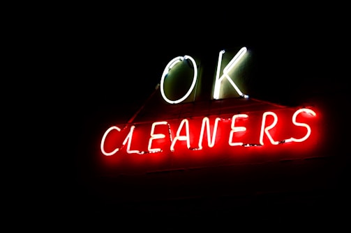 Neon sign displaying the words 'OK CLEANERS' with 'OK' in white and 'CLEANERS' in red letters, glowing brightly against a dark background.