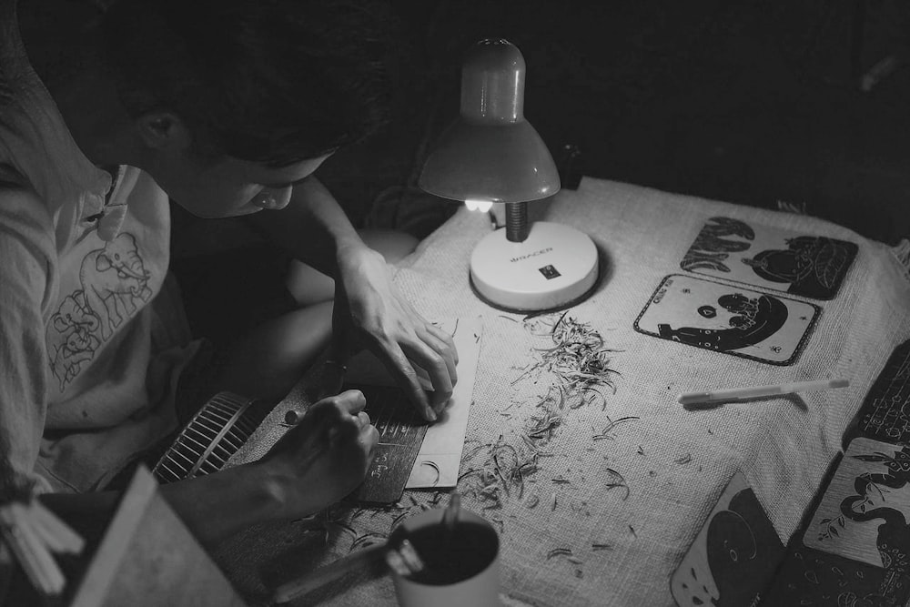 grayscale photography of man writing on brown paper near turned on lamp