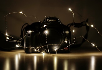 A DSLR camera sits on a reflective surface with a string of small white fairy lights draped around it. The setting is dimly lit, creating a warm glow and highlighting the camera's contours. The focus is on the camera with its brand and model visible.