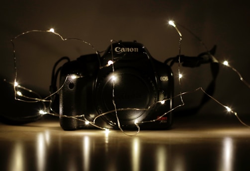A DSLR camera sits on a reflective surface with a string of small white fairy lights draped around it. The setting is dimly lit, creating a warm glow and highlighting the camera's contours. The focus is on the camera with its brand and model visible.