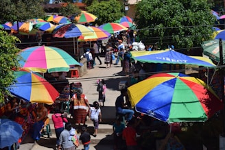 A bustling marketplace filled with large, colorful umbrellas providing shade for the vendors and shoppers. People are seen interacting, walking, and exploring various goods laid out on tables. Vibrant textiles and clothing are prominently displayed under the parasols, while lush green trees add to the lively atmosphere.