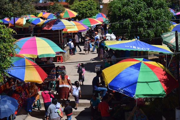 A bustling marketplace filled with large, colorful umbrellas providing shade for the vendors and shoppers. People are seen interacting, walking, and exploring various goods laid out on tables. Vibrant textiles and clothing are prominently displayed under the parasols, while lush green trees add to the lively atmosphere.