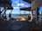An open-air patio overlooking a scenic view of a lake with clear blue skies and distant hills. The seating area includes wicker chairs and round tables, shaded by umbrellas and surrounded by metal railings. The structure features stone walls and industrial design elements.