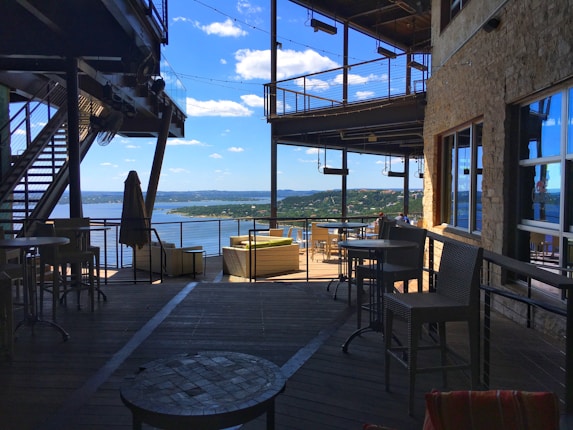 An open-air patio overlooking a scenic view of a lake with clear blue skies and distant hills. The seating area includes wicker chairs and round tables, shaded by umbrellas and surrounded by metal railings. The structure features stone walls and industrial design elements.