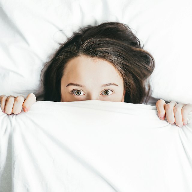 Frau liegt im Bett und zieht die Decke über das Gesicht