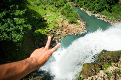 A hand showing a peace sign is extended over a lush green landscape with a waterfall. The water cascades down with force, surrounded by vibrant greenery and rocks. Sunlight illuminates the scene, highlighting the contrast between the water and the surrounding nature.