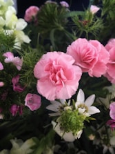 A bouquet of various flowers features prominently, including pink carnations, delicate white flowers, and lush green foliage. The arrangement displays a variety of textures and colors with a natural, fresh appearance.
