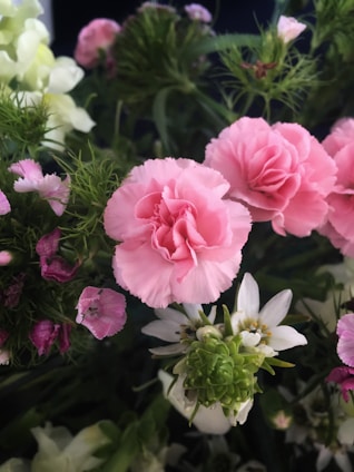 A bouquet of various flowers features prominently, including pink carnations, delicate white flowers, and lush green foliage. The arrangement displays a variety of textures and colors with a natural, fresh appearance.