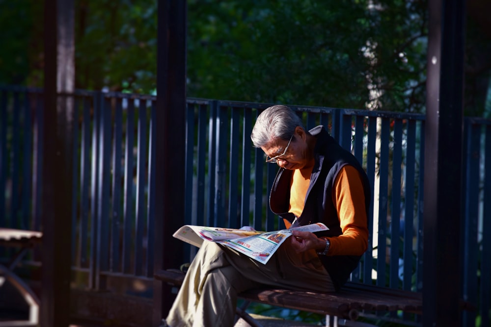 man reading newspaper while sitting on bench in front of wooden fence