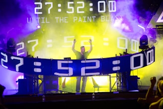 A person stands at a DJ booth surrounded by digital screens displaying a countdown. Bright yellow and purple lights illuminate the area, creating a dynamic, energetic atmosphere. The DJ appears to be engaging with the crowd, raising their hands amidst the haze.