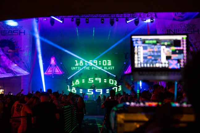 A vibrant concert scene featuring a large digital countdown display on stage, surrounded by colorful lights and energetic crowd. DJ equipment is visible in the foreground, with people dancing and enjoying the event.