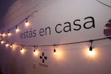 A wall displays the text 'estás en casa' with a string of warm yellow lights hanging across it. The lower part shows the word 'TOLUCA' accompanied by a simple logo. The wall is painted white with a faint shadow effect from the lighting.
