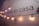 A wall displays the text 'estás en casa' with a string of warm yellow lights hanging across it. The lower part shows the word 'TOLUCA' accompanied by a simple logo. The wall is painted white with a faint shadow effect from the lighting.