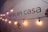 A wall displays the text 'estás en casa' with a string of warm yellow lights hanging across it. The lower part shows the word 'TOLUCA' accompanied by a simple logo. The wall is painted white with a faint shadow effect from the lighting.