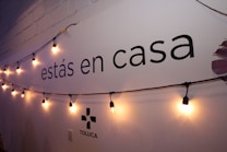 A wall displays the text 'est&aacute;s en casa' with a string of warm yellow lights hanging across it. The lower part shows the word 'TOLUCA' accompanied by a simple logo. The wall is painted white with a faint shadow effect from the lighting.