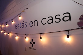 A wall displays the text 'est&aacute;s en casa' with a string of warm yellow lights hanging across it. The lower part shows the word 'TOLUCA' accompanied by a simple logo. The wall is painted white with a faint shadow effect from the lighting.