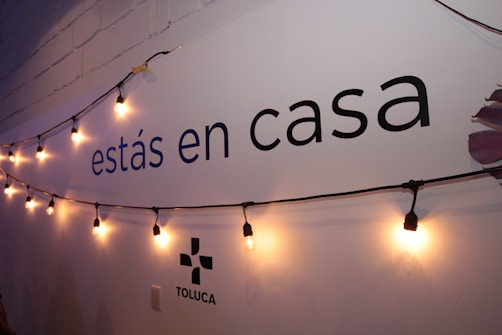 A wall displays the text 'estás en casa' with a string of warm yellow lights hanging across it. The lower part shows the word 'TOLUCA' accompanied by a simple logo. The wall is painted white with a faint shadow effect from the lighting.