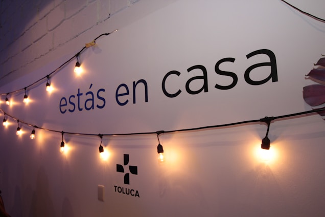 A wall displays the text 'est&aacute;s en casa' with a string of warm yellow lights hanging across it. The lower part shows the word 'TOLUCA' accompanied by a simple logo. The wall is painted white with a faint shadow effect from the lighting.