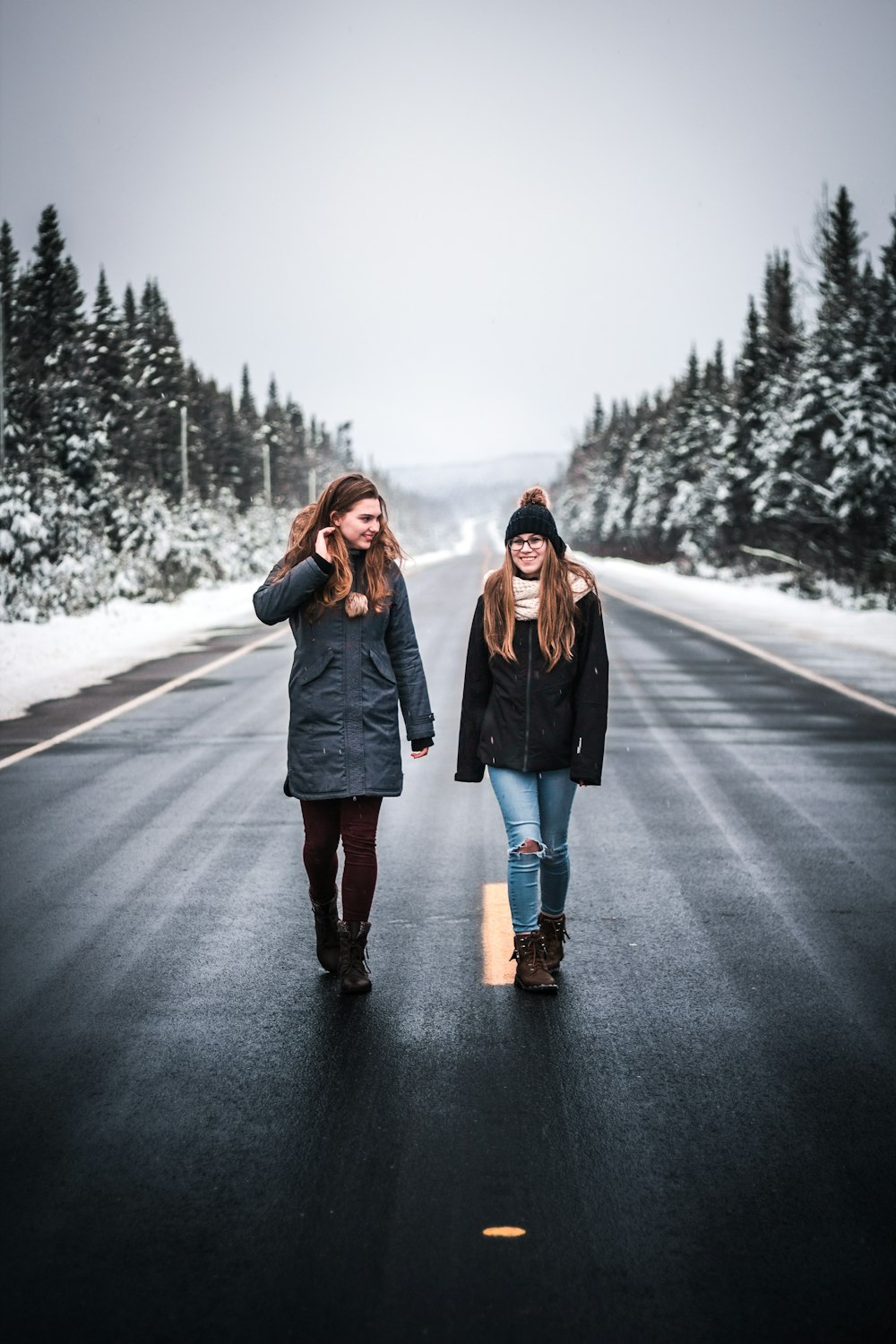 two women walking on gray road between trees