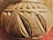 A close-up view of a carved wooden or clay object with intricate leaf and geometric patterns. The texture is rough and appears handmade, with an earthy tone and elaborate design on the surface.