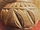 A close-up view of a carved wooden or clay object with intricate leaf and geometric patterns. The texture is rough and appears handmade, with an earthy tone and elaborate design on the surface.