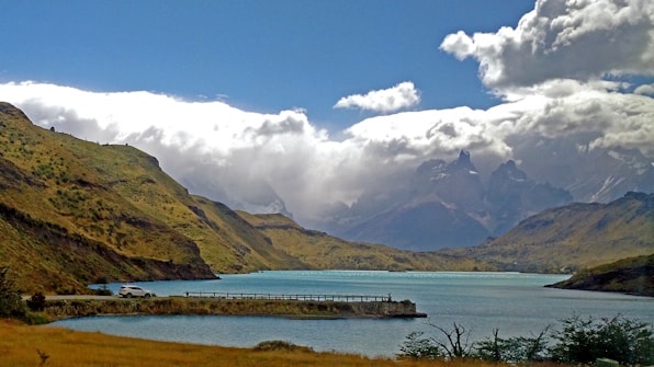 A serene landscape of a calm blue lake surrounded by lush green hills. In the background, majestic mountains are shrouded in dramatic, billowy clouds. A small car is parked by the lakeside road, adding a sense of scale and tranquility.