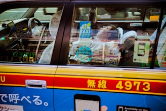 A person is sleeping in a taxi, visible through the window. The interior features lace headrest covers and various signage indicating taxi services. Japanese characters and vibrant colors adorn the exterior of the vehicle, with a prominent blue advertisement on the side.