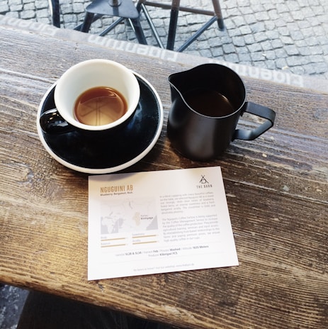 A wooden table holds a black coffee cup on a saucer and a black milk pitcher. Beside them lies a card displaying coffee information, with text and a map. The cobblestone street is visible beyond the edge of the table.