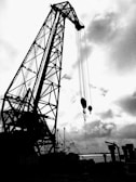 A large industrial crane dominates the scene, silhouetted against a cloudy sky. The structure is intricate with a lattice-like framework and several cables hanging down. In the background, smaller cranes and industrial equipment are visible along a waterfront.