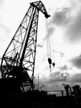 A large industrial crane dominates the scene, silhouetted against a cloudy sky. The structure is intricate with a lattice-like framework and several cables hanging down. In the background, smaller cranes and industrial equipment are visible along a waterfront.