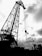 A large industrial crane dominates the scene, silhouetted against a cloudy sky. The structure is intricate with a lattice-like framework and several cables hanging down. In the background, smaller cranes and industrial equipment are visible along a waterfront.