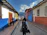 A narrow cobblestone street flanked by colorful colonial-style buildings, with a person walking away carrying a backpack. Another person is standing by the door of a building. The architecture features vibrant blue, red, and white walls with wooden accents, and mountains can be seen in the background under a partly cloudy sky.