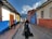 A narrow cobblestone street flanked by colorful colonial-style buildings, with a person walking away carrying a backpack. Another person is standing by the door of a building. The architecture features vibrant blue, red, and white walls with wooden accents, and mountains can be seen in the background under a partly cloudy sky.