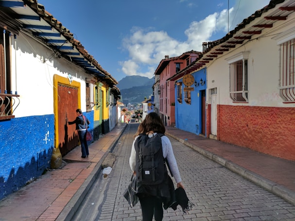 A narrow cobblestone street flanked by colorful colonial-style buildings, with a person walking away carrying a backpack. Another person is standing by the door of a building. The architecture features vibrant blue, red, and white walls with wooden accents, and mountains can be seen in the background under a partly cloudy sky.
