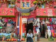 A colorful storefront is filled with a variety of handmade crafts and textiles, including hammocks, bags, blankets, and clothing. The building is adorned with bright decorations and vibrant patterns, with a red facade and green accents. A person is standing in front of the shop, wearing a backpack and observing the display.