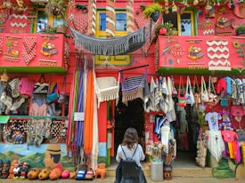 A colorful storefront is filled with a variety of handmade crafts and textiles, including hammocks, bags, blankets, and clothing. The building is adorned with bright decorations and vibrant patterns, with a red facade and green accents. A person is standing in front of the shop, wearing a backpack and observing the display.