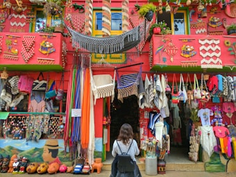 A colorful storefront is filled with a variety of handmade crafts and textiles, including hammocks, bags, blankets, and clothing. The building is adorned with bright decorations and vibrant patterns, with a red facade and green accents. A person is standing in front of the shop, wearing a backpack and observing the display.