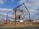 A wooden house structure is suspended at an angle within a large metal frame in an open field. The house appears weathered and aged, with its door partially open. The metal frame consists of four tall, rust-colored beams, supporting the house with wires. The scene is set against a backdrop of dry grass and sparse trees under a partly cloudy sky.