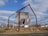 A wooden house structure is suspended at an angle within a large metal frame in an open field. The house appears weathered and aged, with its door partially open. The metal frame consists of four tall, rust-colored beams, supporting the house with wires. The scene is set against a backdrop of dry grass and sparse trees under a partly cloudy sky.
