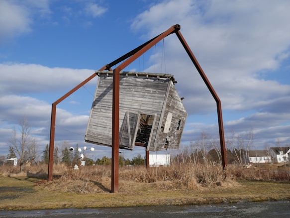 A wooden house structure is suspended at an angle within a large metal frame in an open field. The house appears weathered and aged, with its door partially open. The metal frame consists of four tall, rust-colored beams, supporting the house with wires. The scene is set against a backdrop of dry grass and sparse trees under a partly cloudy sky.