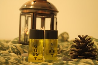 Two small bottles labeled 'guest shampoo' are placed alongside a pinecone and in front of a bronze lantern with star-shaped cutouts. The setting includes a green fabric with a camouflage pattern and a few stones scattered around, creating a rustic and cozy atmosphere.