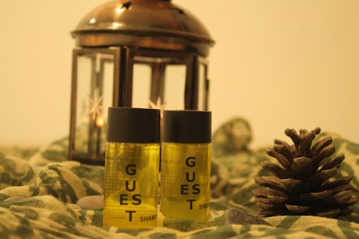 Two small bottles labeled 'guest shampoo' are placed alongside a pinecone and in front of a bronze lantern with star-shaped cutouts. The setting includes a green fabric with a camouflage pattern and a few stones scattered around, creating a rustic and cozy atmosphere.