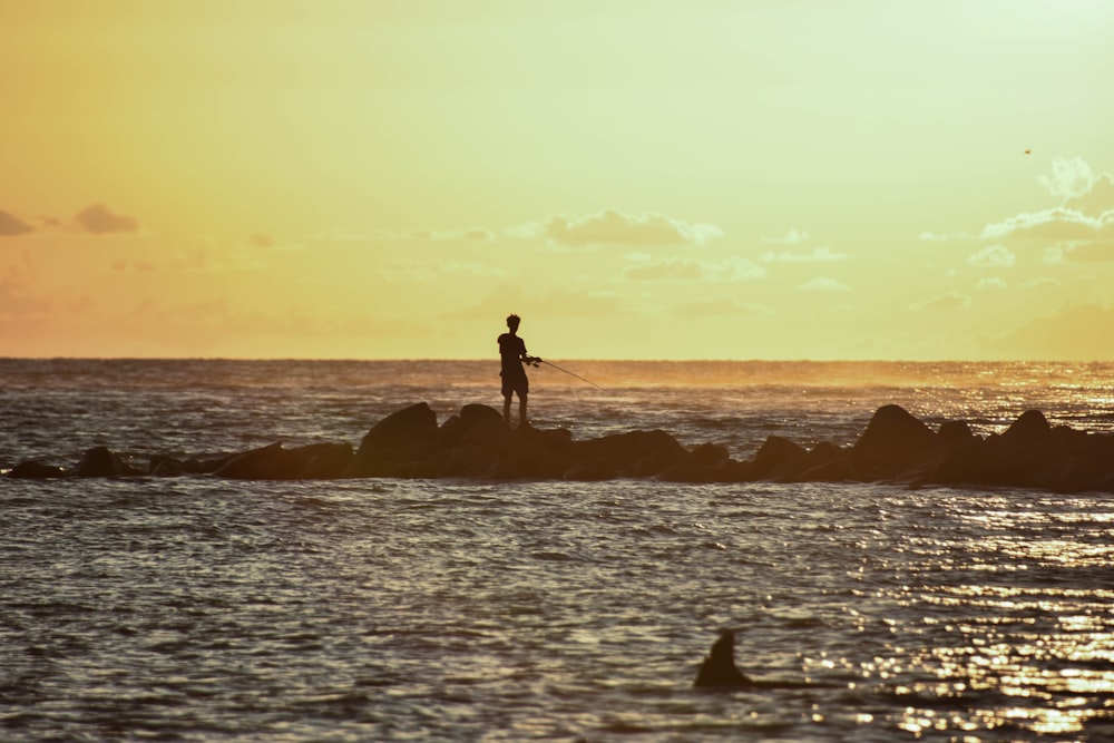 silhouette of person standing on rock formation surrounded by body of water during sunset