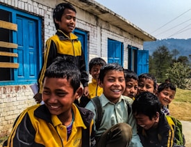 A group of smiling boys in yellow and black uniforms are gathered outside a brick building with blue doors. They appear to be in a joyful and playful mood, situated in a rural setting with trees and hills in the background.