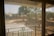 A view through a window screen showing an outdoor scene with blurred trees and landscape. The mesh of the screen creates a grid-like pattern, with the light filtering through it.